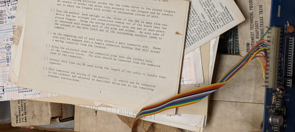 a pile of assorted papers and schematics resting on top of a PCB with a rainbow ribbon cable protruding